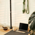 A yoga mat on a wooden floor in a bright room, styled with an open laptop, a small reed diffuser, a glass of water, a bowl, and a tall black bar. A green hanging rope is visible on the wall, along with a blurred plant in the foreground.